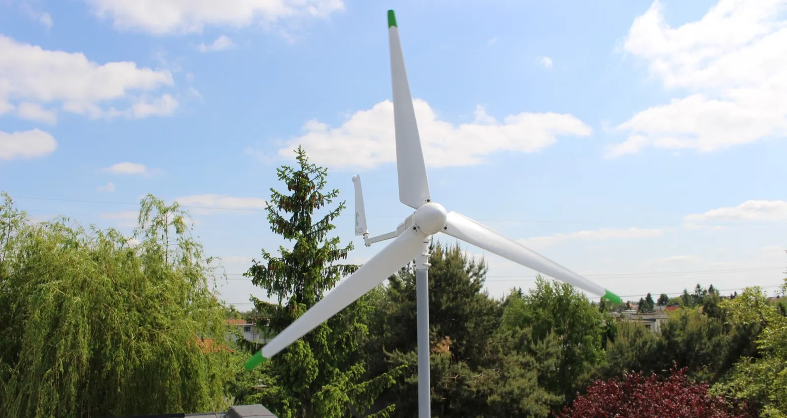 Mała pozioma turbina wiatrowa na maszcie przy domu, w tle drzewa i niebo